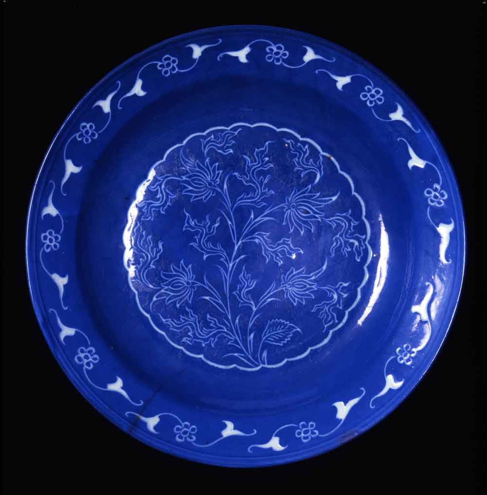 Ceramic dish with Chinese-inspired flower, Iran, Kirman?; 17th century Innumerable copies of blue and white Chinese export porcelain were made in Iran from the 15th to the 17th century. The quality varied, and only rarely did the results have independent artistic value. This magnificent dish does, its design created by the potter cutting through the intense blue slip down to the white fritware. While the simple pattern on the rim, consisting of double split leaves joined by a five-petaled flower, shows ties to Islam, the nervously quivering plant in the center is oriented toward China. The plant has four lotus-like flowers, a few simple little leaves, and other more eccentric ones that resemble the jagged cloud ornaments that surround or perhaps emerge from the flowers.
