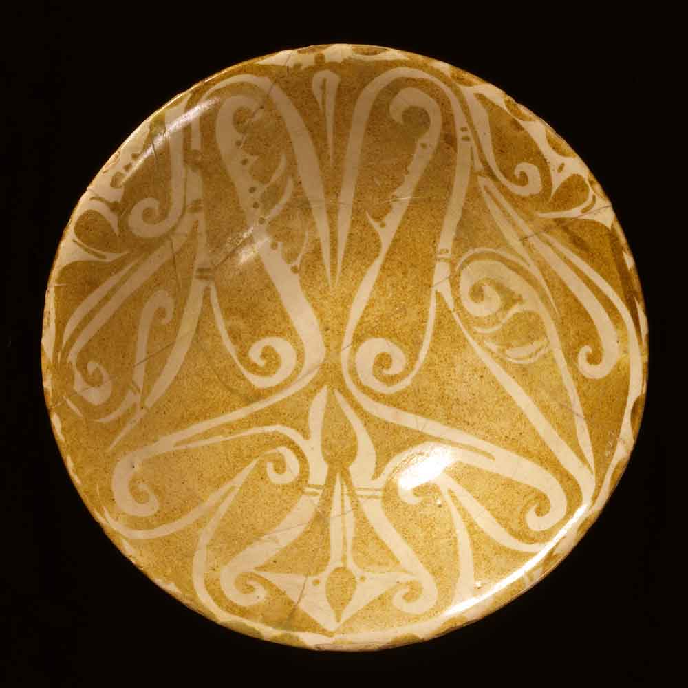 Ceramic bowl with luster decoration<br />Iraq; 9th-10th century<br />The beveled style is three-dimensional almost by definition, but is also found in two-dimensional versions, as on this unusual bowl decorated in luster.<br />The design consists largely of halfpalmettes, some of which are highly stylized while others are laciniated and slightly more naturalistic. As on arabesques, they grow from or extend one another unnaturally. It is difficult to differentiate between background and pattern, however, on closer inspection, it becomes clear that the golden luster decoration is what forms the complex palmette motif.