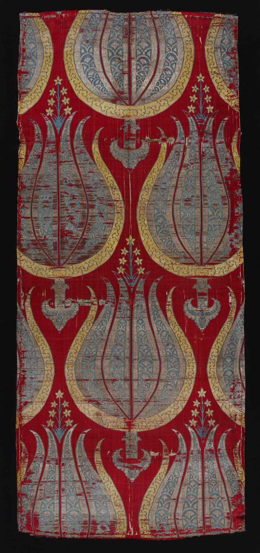 Silk with stylized tulips, Turkey; 2nd half of 16th century <br><br> The designs of large-patterned Ottoman silks from the 16th century are among the most sublime woven anywhere. They were made in Bursa and Istanbul, and their patterns were often designed by the artists in the court studio (nakkaşhane). The most impressive group of these fabrics can in fact be found in the Topkapi Palace’s collection of the Ottoman sultans’ caftans. <br> Rows of stylized tulips form the pattern. The tulips have six petals: the outermost two are golden and blue with Chinese cloud ornaments, and the four innermost are silver and blue with a scale pattern. A seven-headed stamen and an odd stalk further denaturalize the motif. The use of precious metals for these textiles resulted in several bans on weaving them.