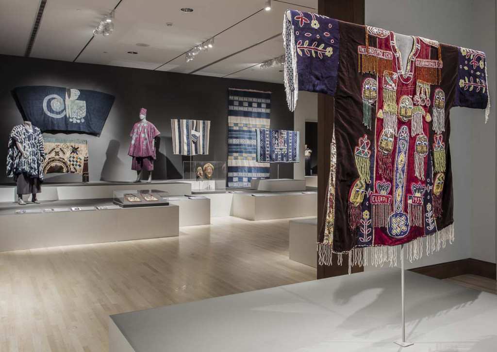 Gallery installation of 'Majestic African Textiles' at Indianapolis Museum of Art showing various West African narrow stripe woven cloths to the right, a Hausa agbaba robe on the wall left and a N'dop Cameroonian costume front left and in the foreground a Yoruba ceremonial tunic from the early 20th century 