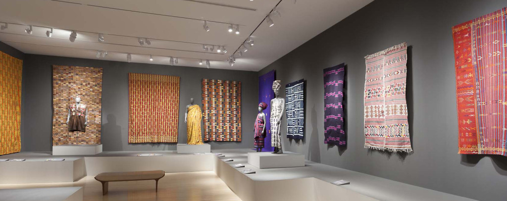Gallery installation of 'Majestic African Textiles' at Indianapolis Museum of Art showing various Ewe and Kente narrow strip woven cloths, alongside Cameroonian and Nigerian cloths 