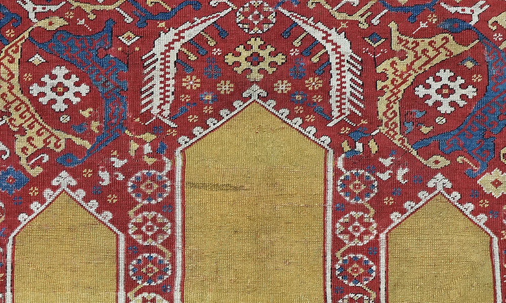 The richly coloured and decorated spandrels above the three-part yellowground niche in this ‘coupled-column’ prayer rug (the term was first used by May H. Beattie in an article of the same name published in Oriental Art, XIV/4, 1968, pp. 243-258), are filled with tw0 varieties of paired curved ragged leaf forms enclosing snowflake rosettes. Those in white create an implied additional gable above the central broad yellow niche, while the blue and yellow Ottoman saz leaves above the lower and slightly narrower f lanking subsidiary niches curl back to touch each other, enclosing the rosettes in a manner reminiscent of the herati design seen in Persian carpets. In both instances the stylisation is such that the leaf motifs can be seen as being zoomorphic in nature, perhaps dragon forms. Carefully planned eight-pointed star rosettes and arabesques complete the internal decoration of this elaborate but wellbalanced panel